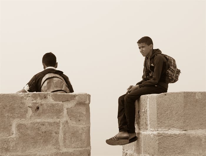 Two boys with backpacks sitting on separate stone walls against a plain sky, sepia