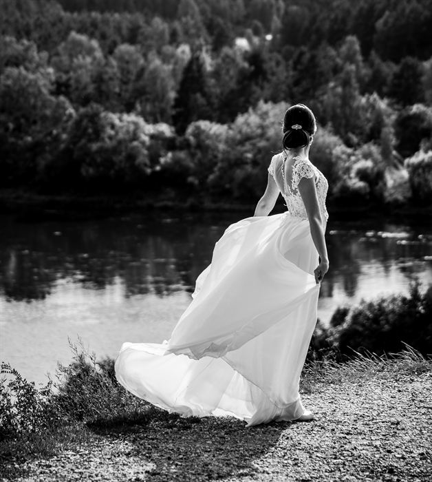 A bride seen from behind, her skirt swinging, on a riverbank, black and white
