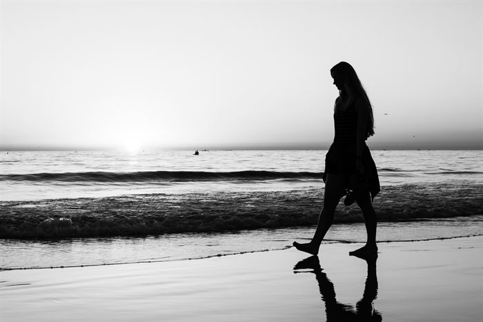 A woman walking along the waterline in silhouette against a low sun, black and white