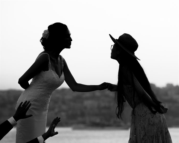 A bride greeting a guest in a wide-brimmed hat, both in silhouette, black and white