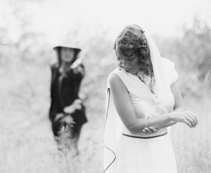 A bride adjusting her bracelet in a field, a guest out of focus behind, black and white