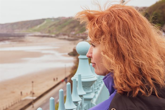 A red-haired woman looking out over a sandy bay from a painted iron railing, England
