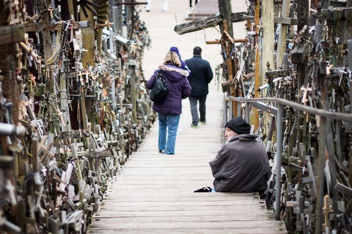 People walking a boardwalk between dense walls of crosses, a man sitting at the side, Hill of Crosses, Lithuania