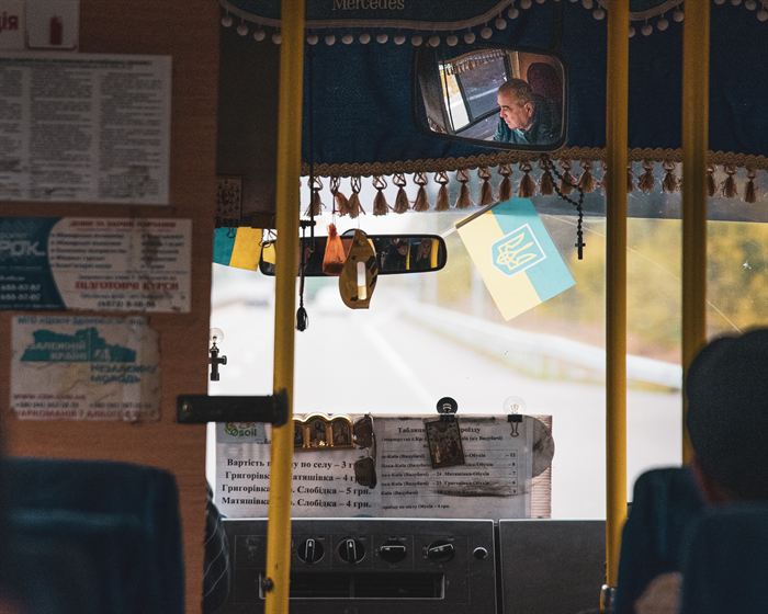 Interior of a rural minibus with a Ukrainian flag, a rosary and a printed fare list, the driver visible in the rear-view mirror