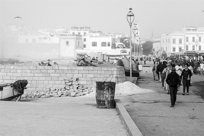 People walking a hazy seafront promenade beside the white ramparts of a Moroccan coastal town, black and white