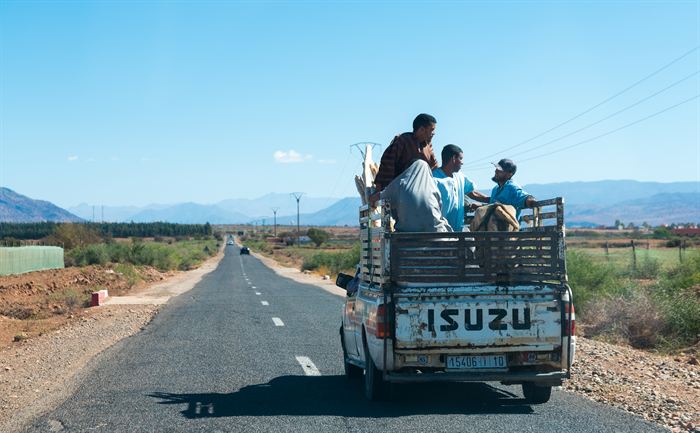 Three men riding in the open bed of a pickup truck on a straight road across arid mountains in Morocco