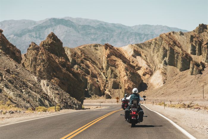 A motorcyclist on an empty highway curving between striped desert rock formations