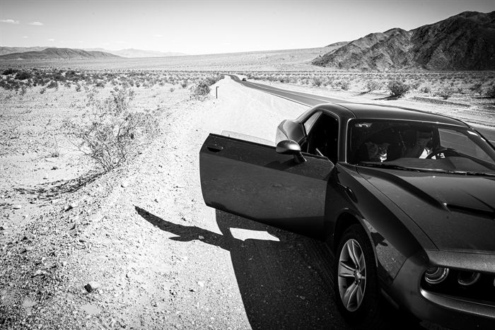 A car stopped on a desert roadside with the driver door open, black and white