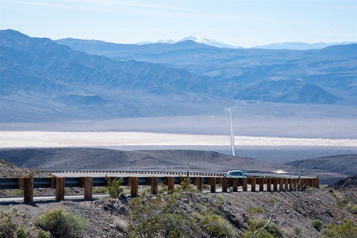 A road descending towards a dry salt flat, a car passing behind a wooden crash barrier