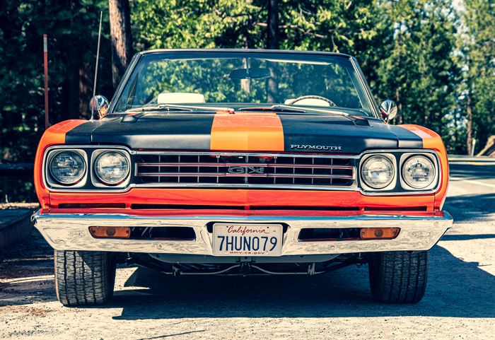 The front of an orange and black Plymouth GTX parked among pine trees