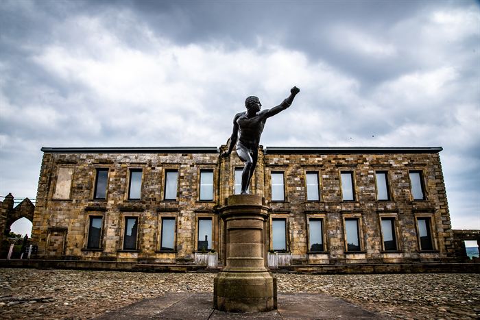 A bronze statue with one arm raised in front of a weathered stone building under heavy cloud