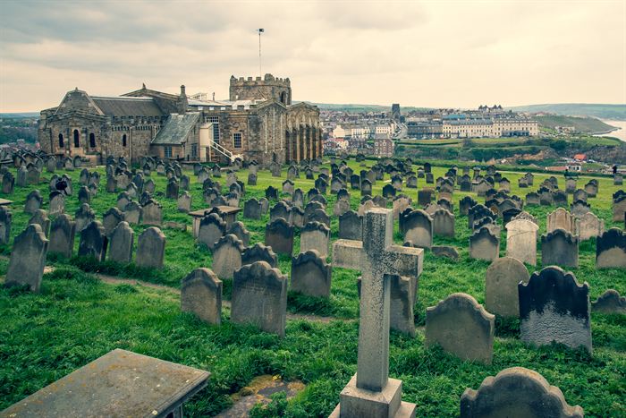 A hillside churchyard of leaning gravestones with an abbey church and the sea behind, Whitby